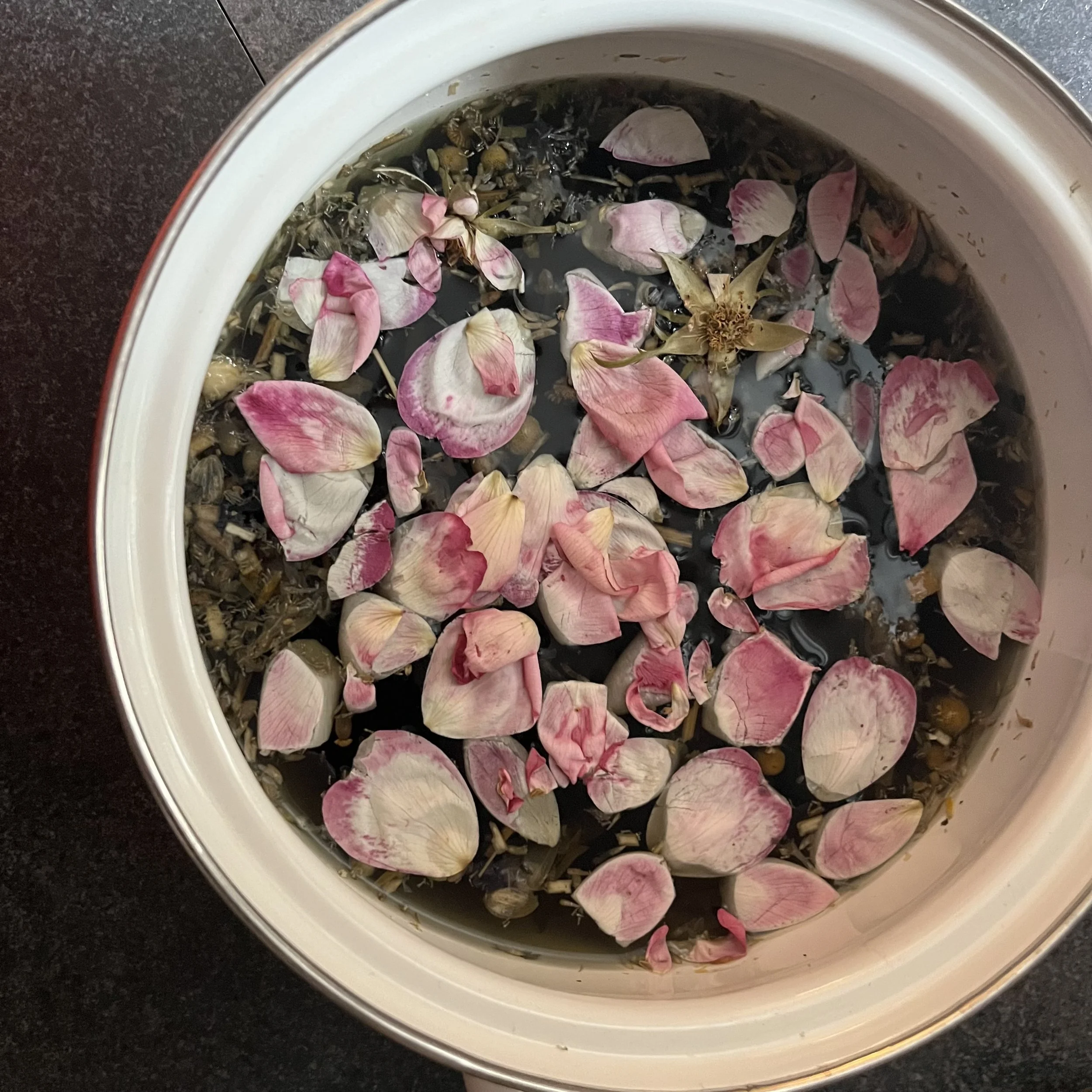 A slow cooker filled with water, pink and white rose petals, and dried herbs.