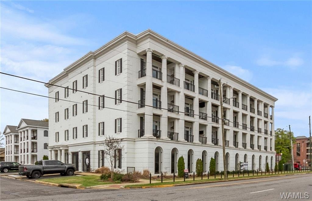 Large white multi-story apartment building with columns and balconies, surrounded by small trees and parked cars, under a blue sky.