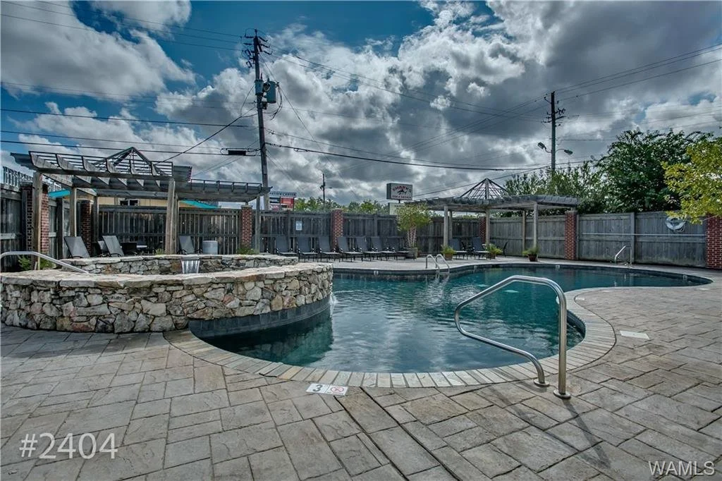 Empty outdoor swimming pool with a hot tub, lounge chairs, and shaded areas on a cloudy day surrounded by a wooden fence.