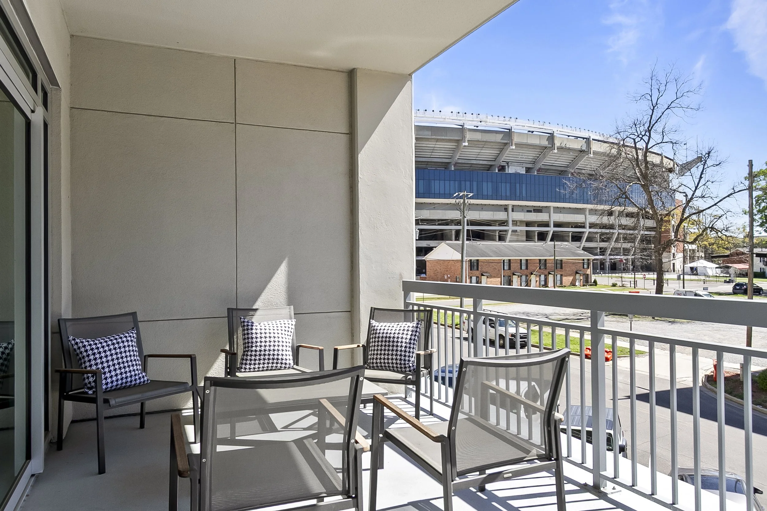 A balcony with four black metal chairs with cushions, overlooking a parking lot with cars, and a large stadium in the background under a clear blue sky.
