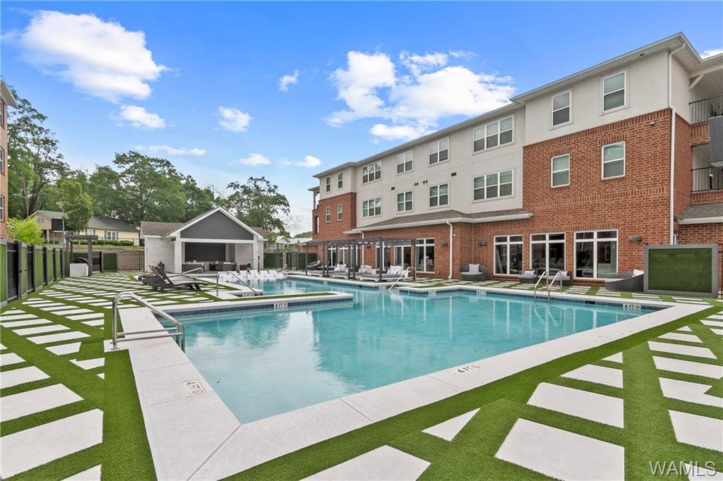 An outdoor swimming pool area in a residential complex with a multi-story apartment building, poolside seating, a small covered shelter, and a clear sky with some clouds.