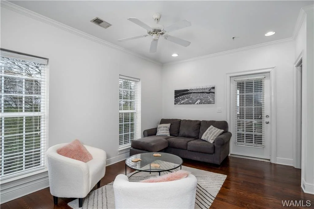 A living room with white walls, hardwood floors, a gray sofa with pillows, two white armchairs with pink pillows, a round glass coffee table, a black and white wall art, windows with blinds, and a door with blinds leading outside.