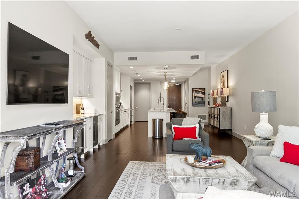 Open-concept living room with gray and white furniture, a large flat-screen TV, a rustic shelving unit, a kitchen with an island, white cabinets, and hardwood floors.