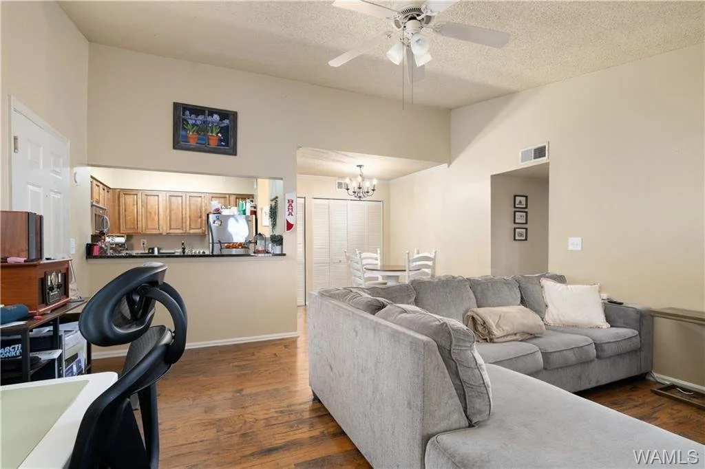 Living room with grey sectional sofa, wooden floors, and a ceiling fan, opening to a kitchen and dining area with white chairs and a chandelier.