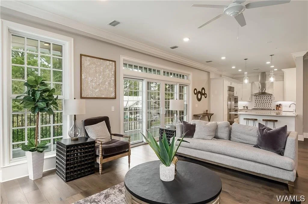 Living room with large windows, a light-colored sofa, a dark armchair with a pillow, a round coffee table with a plant, a potted plant, and an open kitchen in the background.