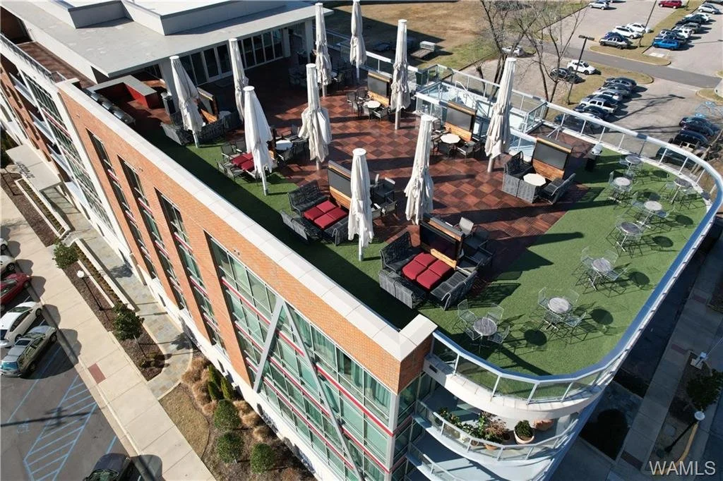 A rooftop balcony on a multi-story building with outdoor seating, umbrellas, and tables, overlooking a parking lot and neighboring area.
