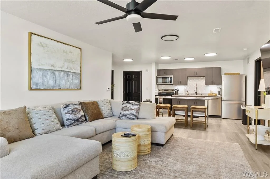 Living room with a large light-colored sectional sofa, patterned throw pillows, and two round wooden coffee tables, adjacent to an open kitchen with gray cabinets, a stainless steel refrigerator, and bar stools.