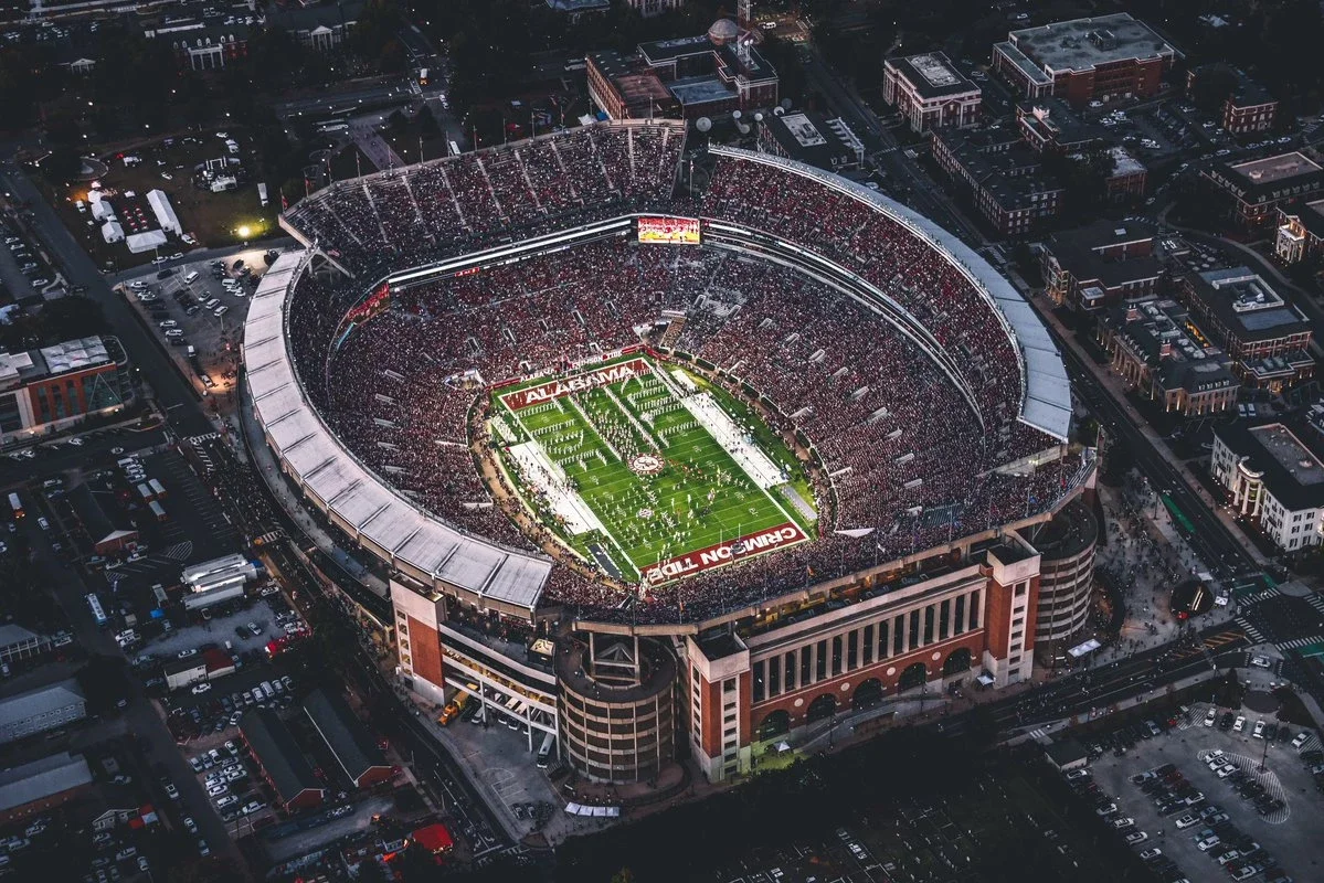 Aerial view of a packed football stadium during a game, surrounded by city buildings at dusk.