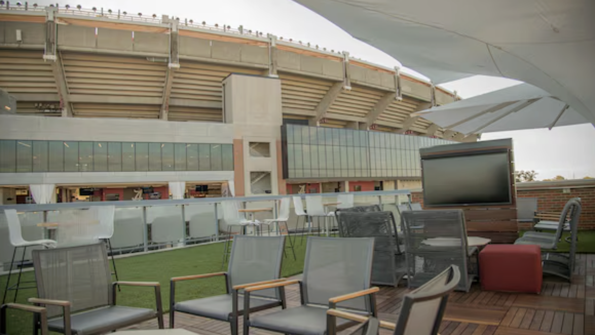 An outdoor rooftop lounge area with chairs and a large flat-screen TV, overlooking a stadium with a modern architectural design.