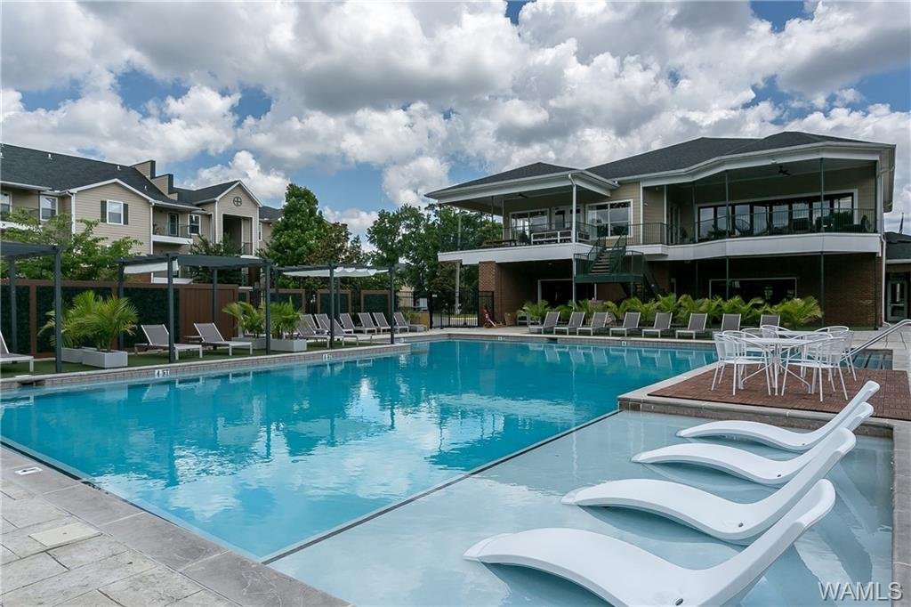 Outdoor swimming pool with lounge chairs and umbrellas, surrounded by residential buildings and a clubhouse with a balcony, under a partly cloudy sky.
