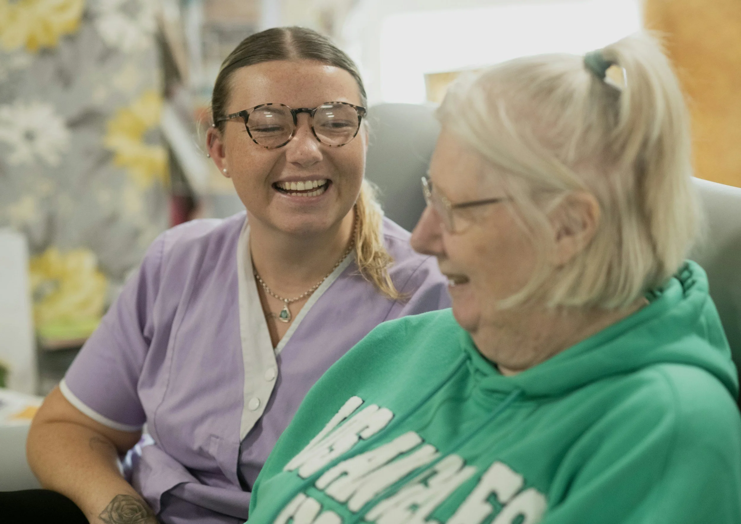 A young woman with glasses and a nursing uniform smiling and talking to an elderly woman with glasses and a green hoodie.