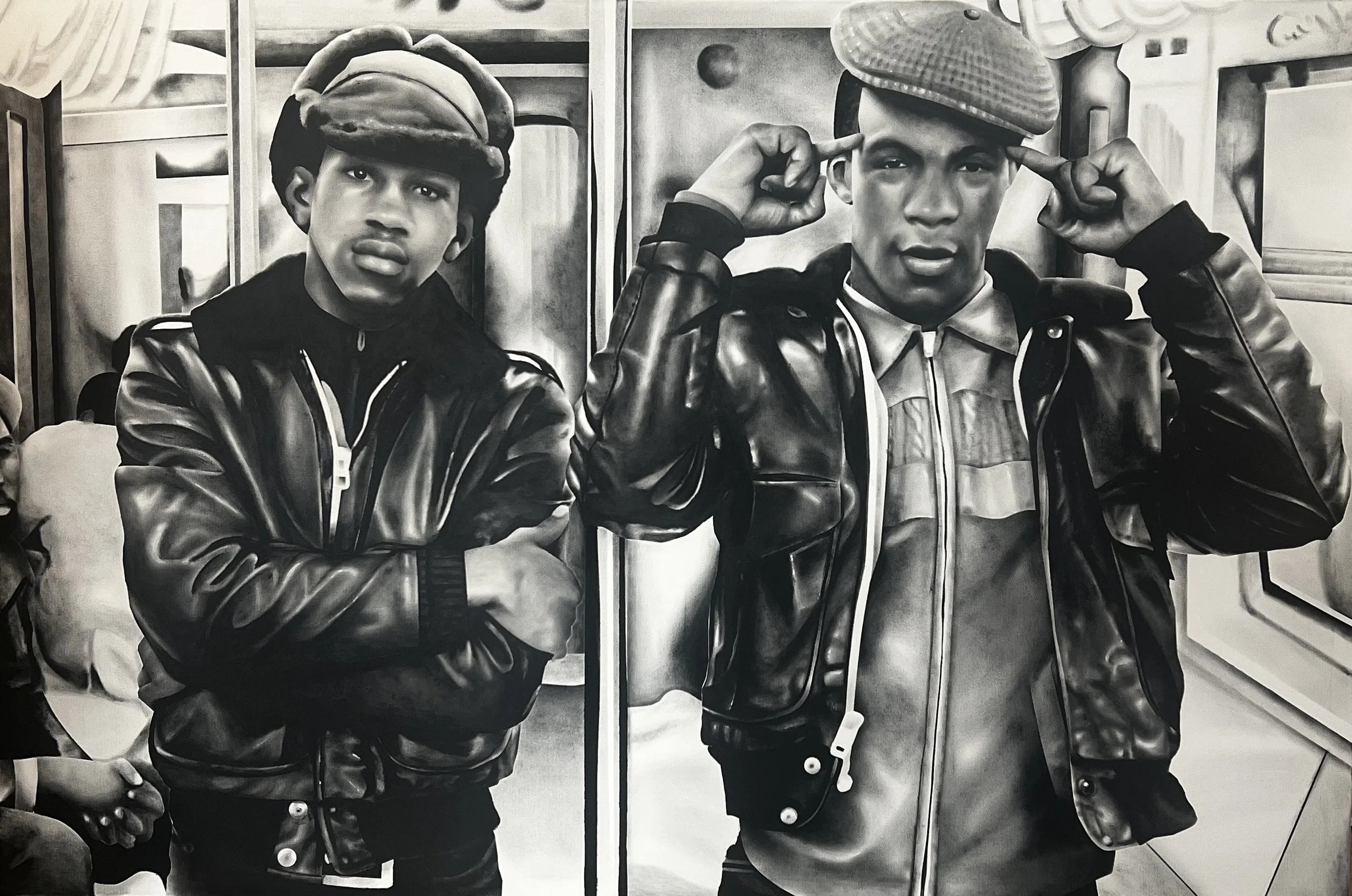 Two young men wearing leather jackets and caps, standing indoors with a background of wooden cabinets and a stove. One has his arms crossed, and the other is touching his cap with both hands.