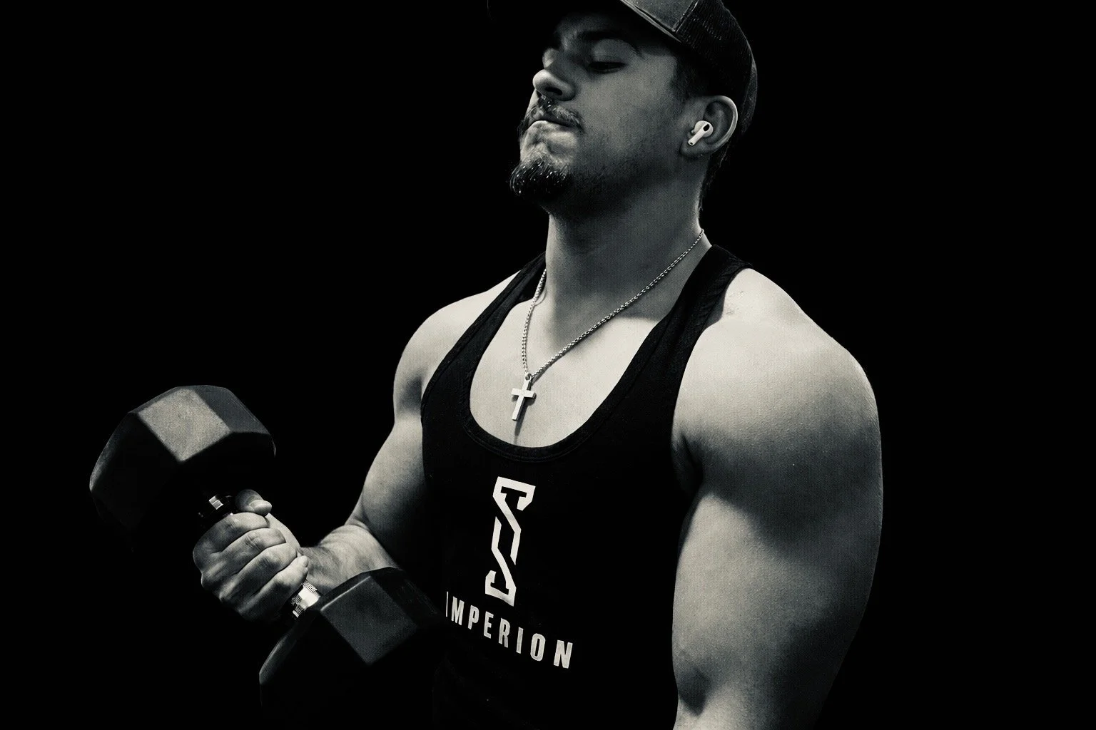 A muscular man lifting a dumbbell, wearing a black tank top with a logo that says "IMPERION," a baseball cap, necklace with a cross pendant, and wireless earbuds, against a black background.