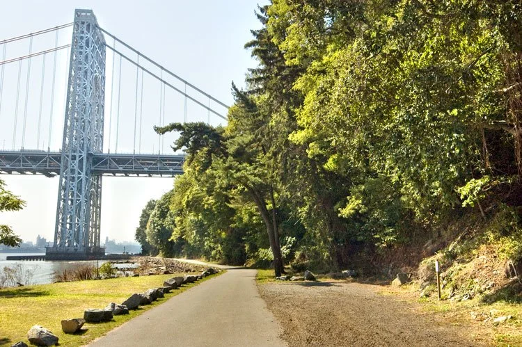 A pathway alongside a river with a suspension bridge in the background. Lush green trees line the right side of the path, casting shadows on the ground.