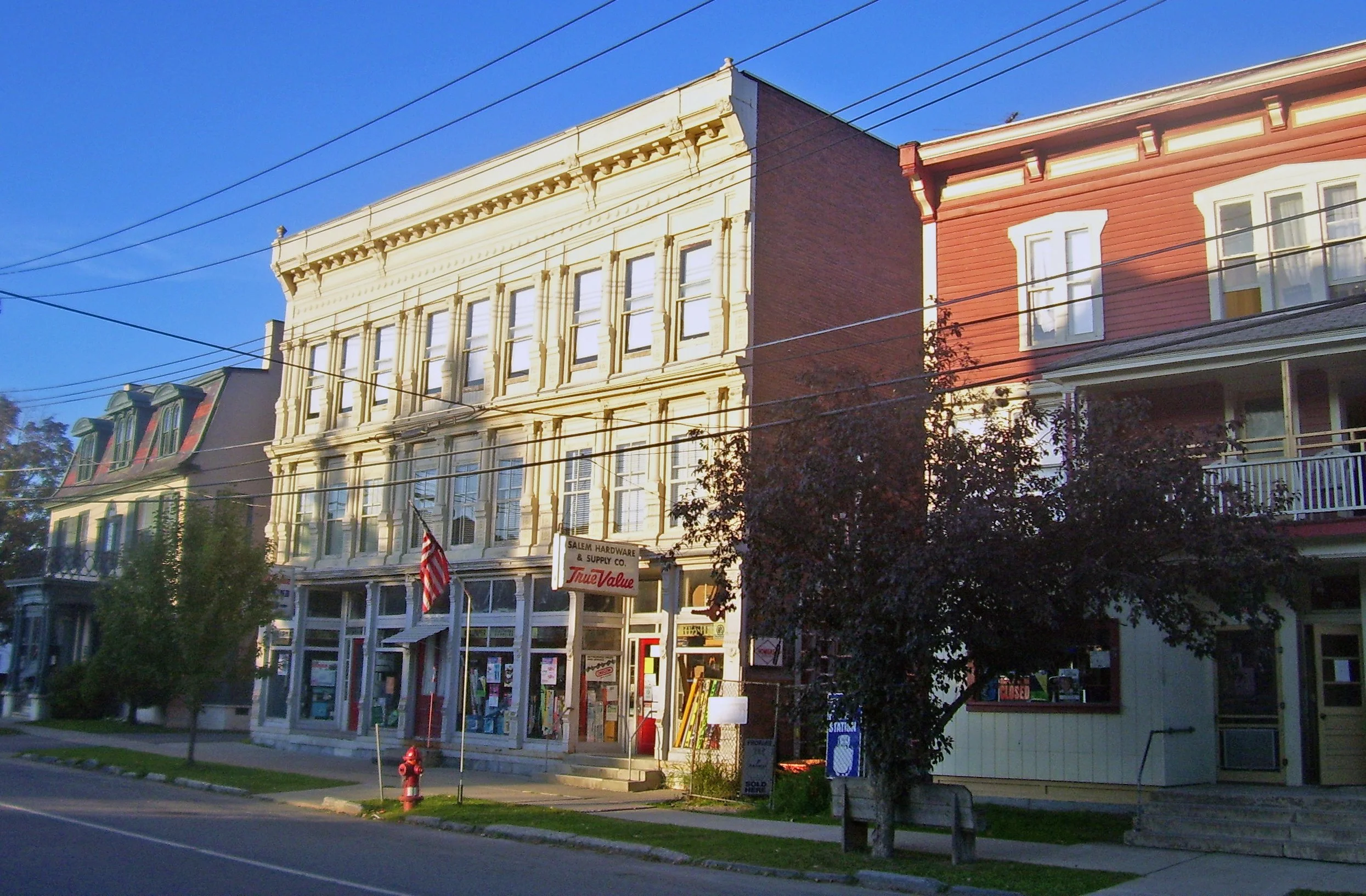 Street view of historic buildings with storefronts, trees, and utility wires, under a clear blue sky in a small town.