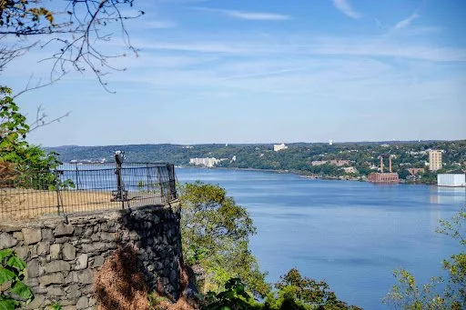 A scenic view of a river or lake with a stone viewing platform and black metal railing, surrounded by greenery, with a distant city skyline and hills under a partly cloudy sky.