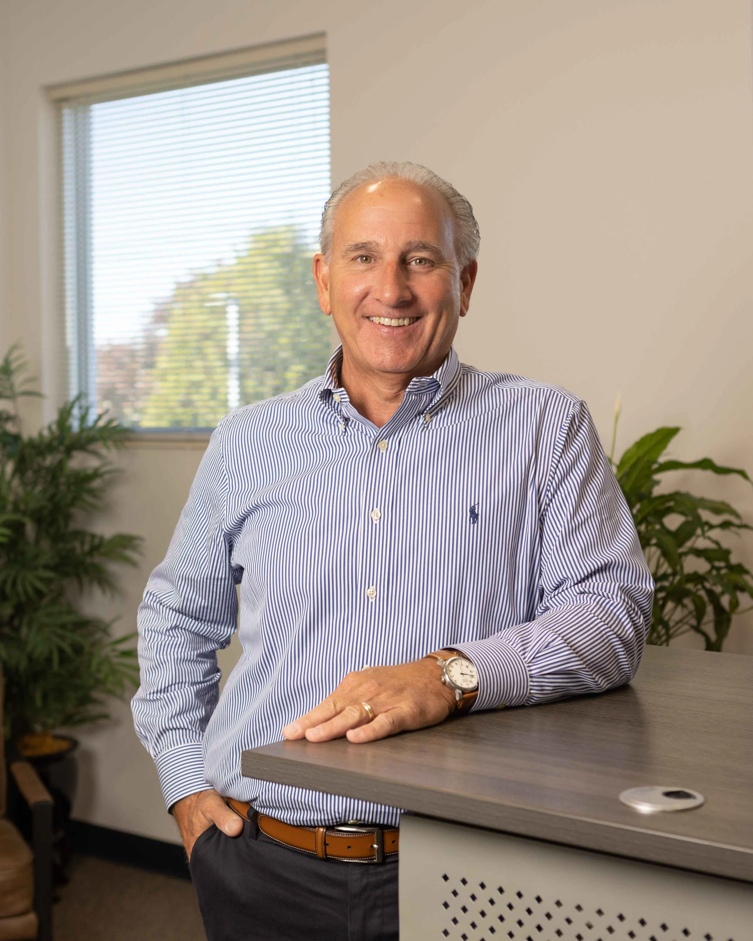 A smiling middle-aged man in a striped button-down shirt and watch, standing in an office with plants and a window.