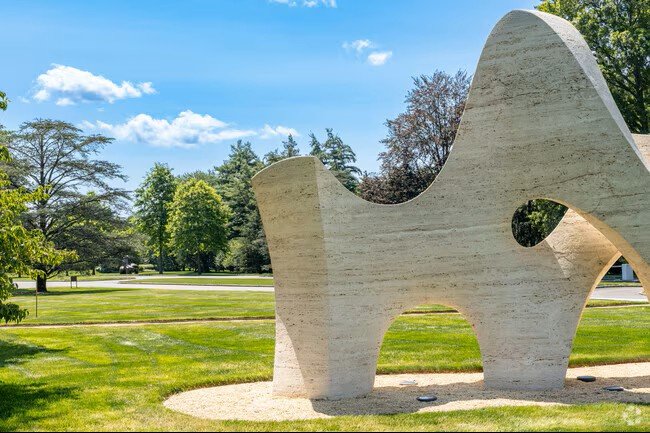 Modern abstract stone sculpture on a grassy lawn in a park with trees and a blue sky.