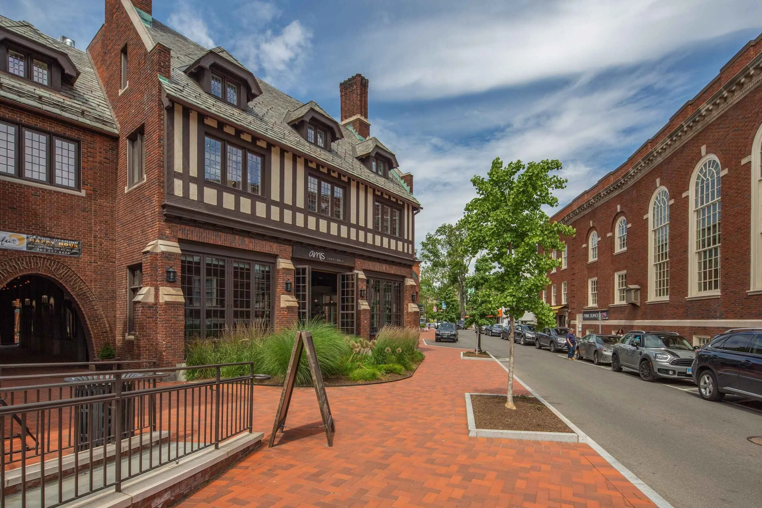 A street scene with brick buildings, trees, parked cars, and a brick-paved sidewalk under a partly cloudy sky.