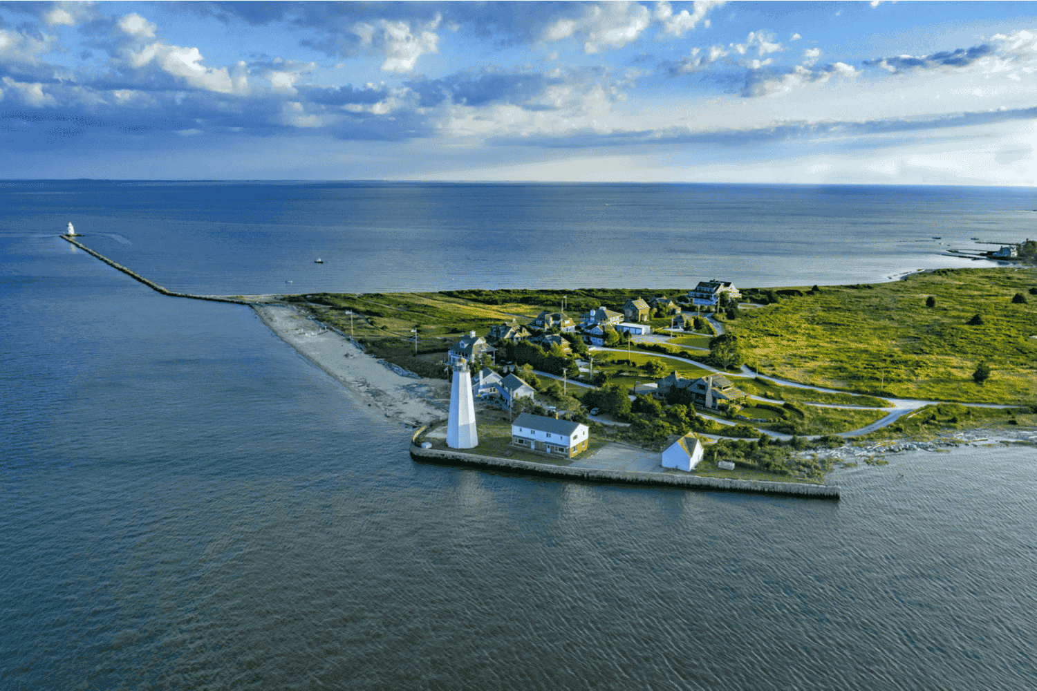 Aerial view of a coastal area with a lighthouse, houses, and a breakwater extending into the ocean, with green land and a partly cloudy sky in the background.