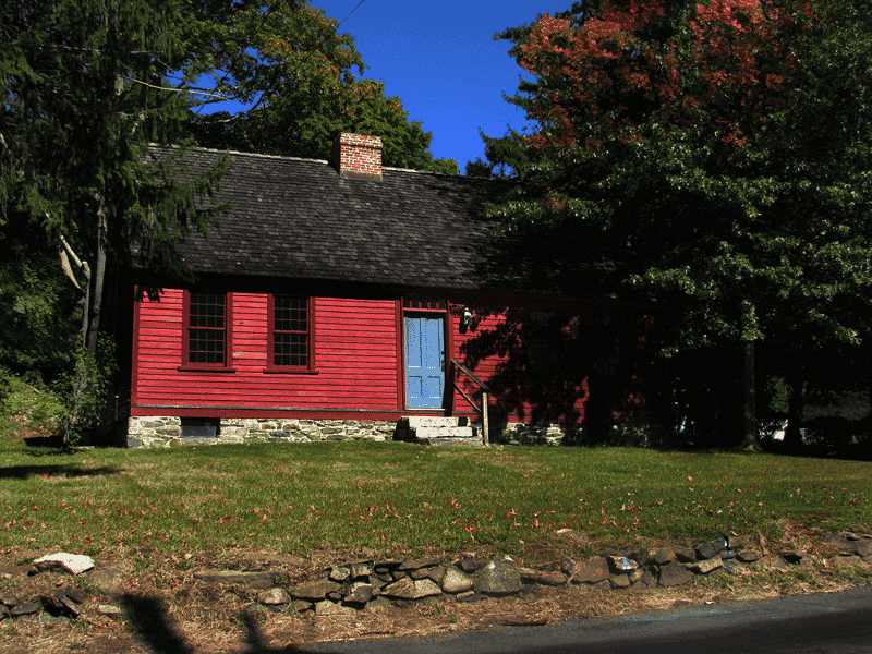 A small, red wooden house with a stone foundation and a blue door, surrounded by trees with green and red leaves, under a bright blue sky.