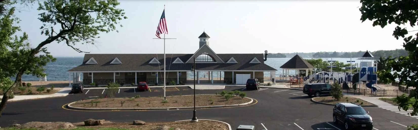 A waterfront building with a parking lot in front, an American flag on a pole, and a playground area to the right, overlooking a body of water.
