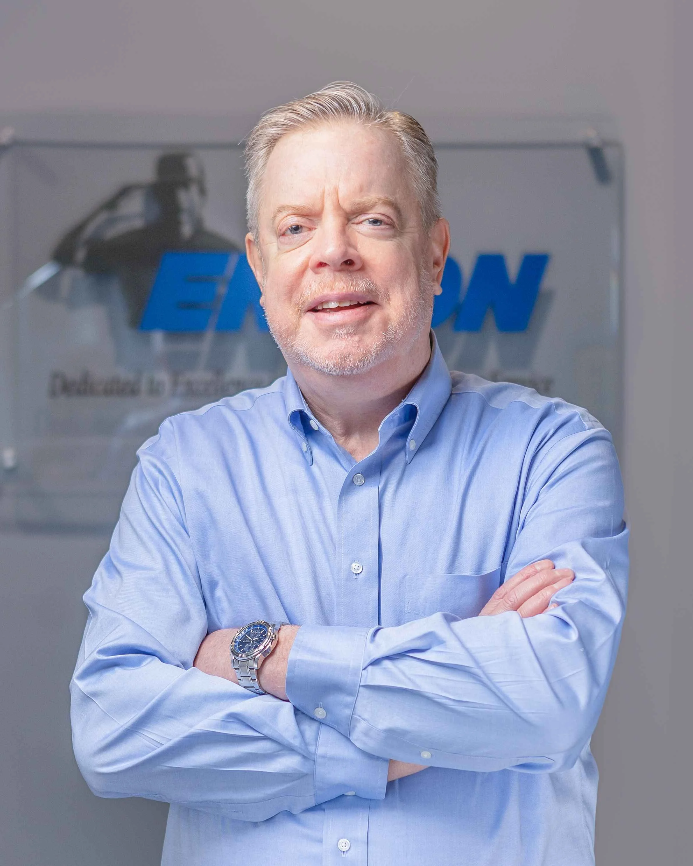 A middle-aged man with light hair and a beard, wearing a blue dress shirt, standing with crossed arms in front of a sign that reads 'ENON' in a professional office setting.