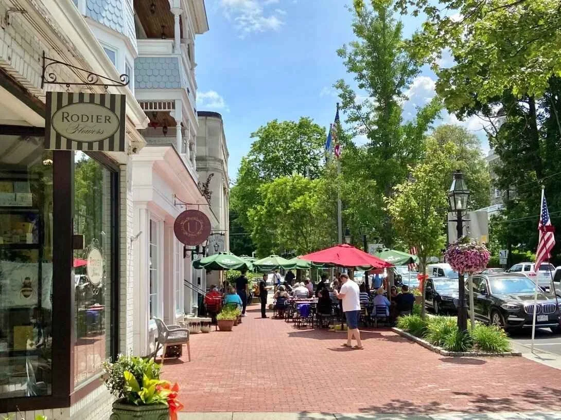 Sidewalk cafes with green and red umbrellas, people sitting and walking, trees, parked cars, and storefronts on a bright, sunny day.