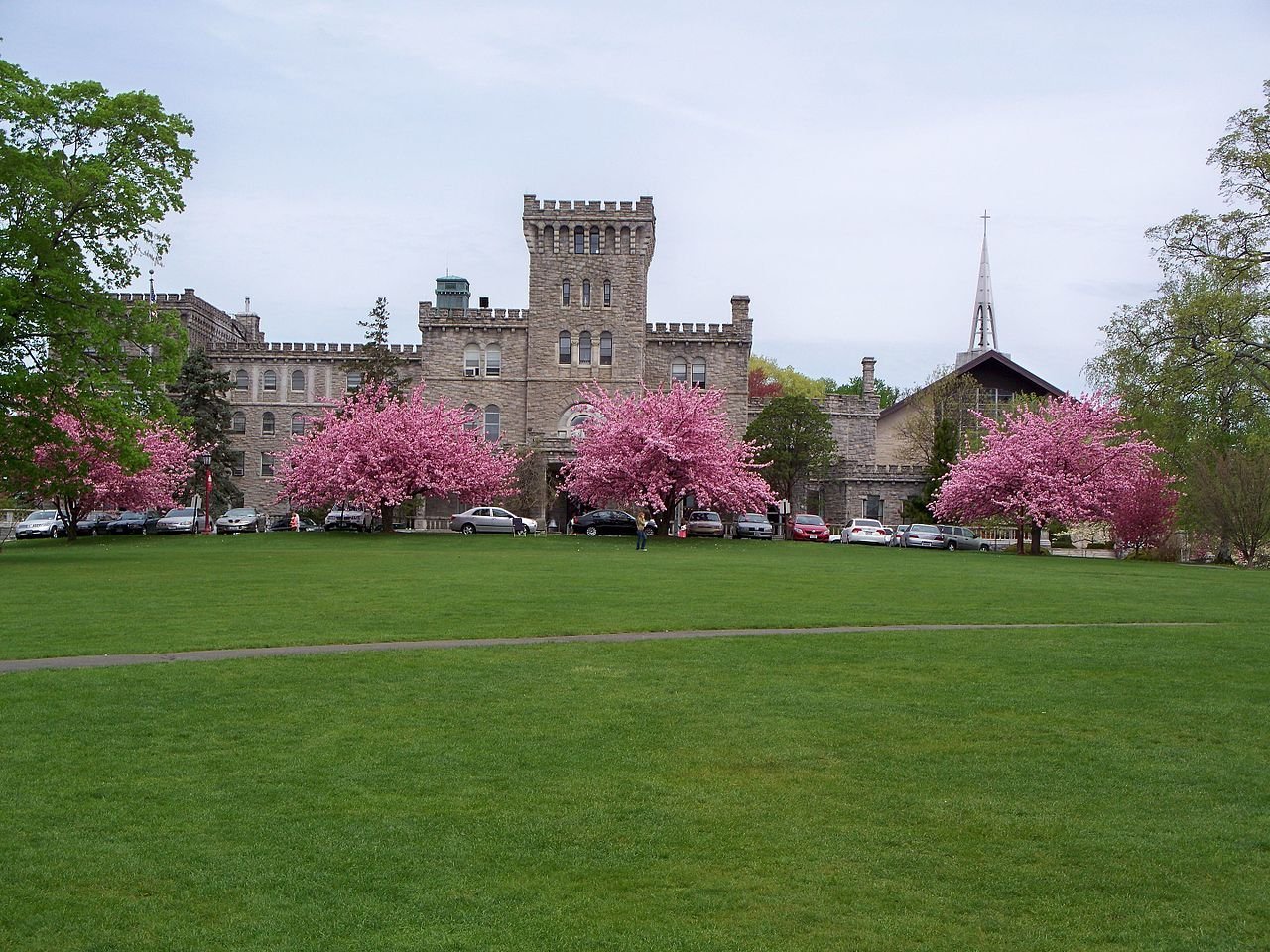 Castle-like stone building with pink blossoming trees in front and parked cars along the front