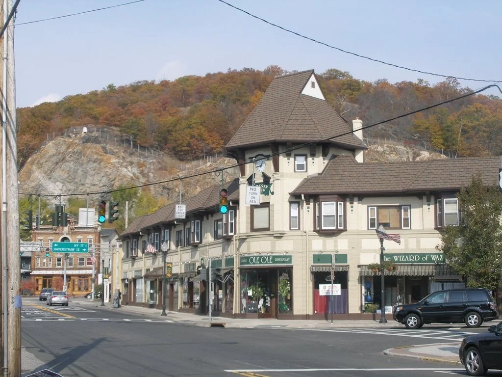 A street scene in a small town with a large building labeled 'Ole Ole' and 'The Wizard of Waz' at the corner, with colorful trees on a hill in the background.