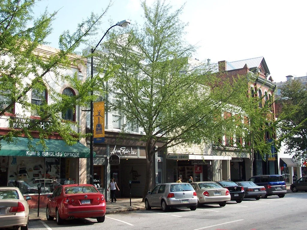 Street view of shops and parked cars on a city street with trees, lamp posts, and buildings in the background.