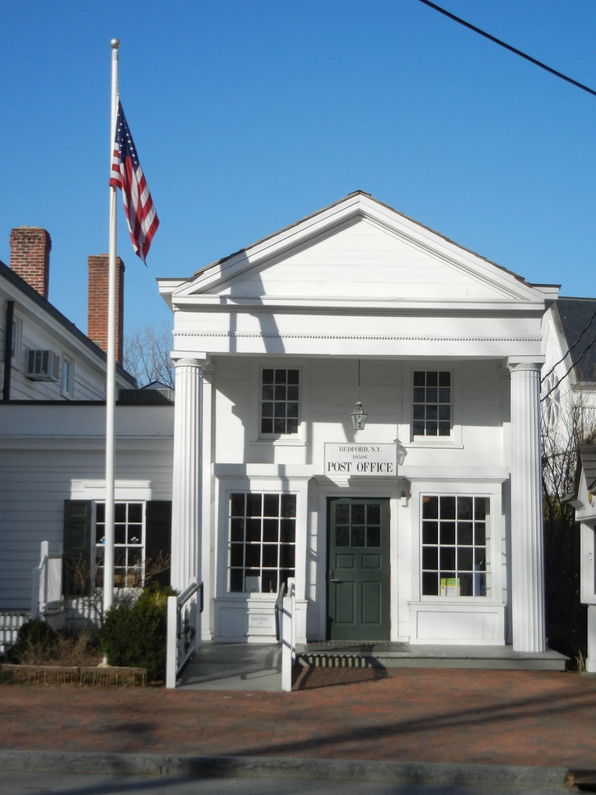 White historic post office building with columns and an American flag flying on a flagpole in front, clear blue sky.