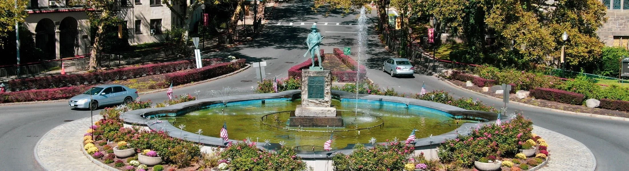 A roundabout with a fountain featuring a statue of a historical figure in the center, surrounded by blooming flowers and small American flags, with cars driving around it.