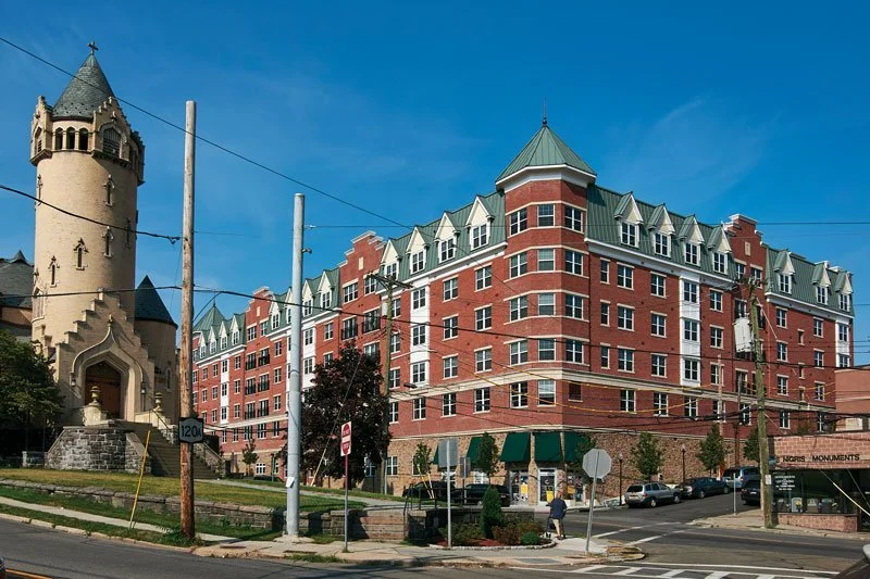 Red brick multi-story building with green rooftops and dormer windows, next to a beige castle-like structure with a tower, under a blue sky in an urban setting.