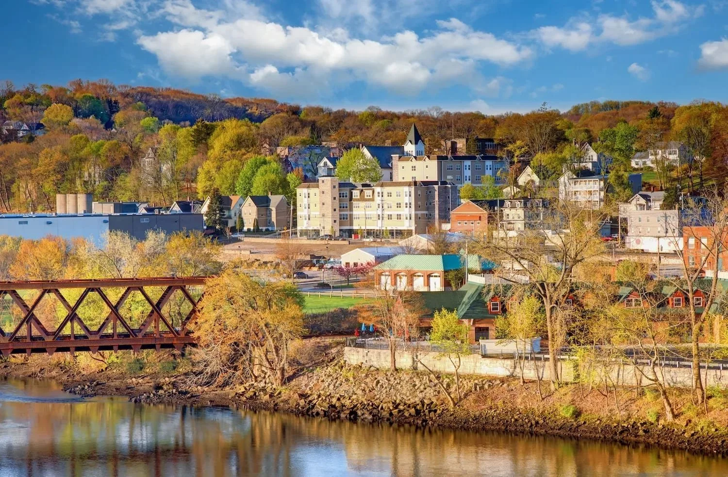 A cityscape with colorful autumn trees, residential buildings, a bridge over a river, and a hill in the background under a partly cloudy sky.