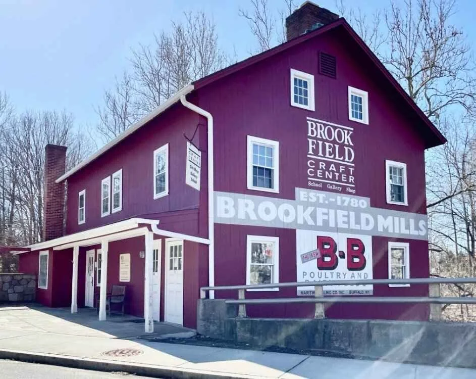 A three-story purple building with white trim advertising Brookfield Mills, a craft center established in 1780, featuring signs for a school, gallery, and shop, with a large red and black B&B Poultry sign.