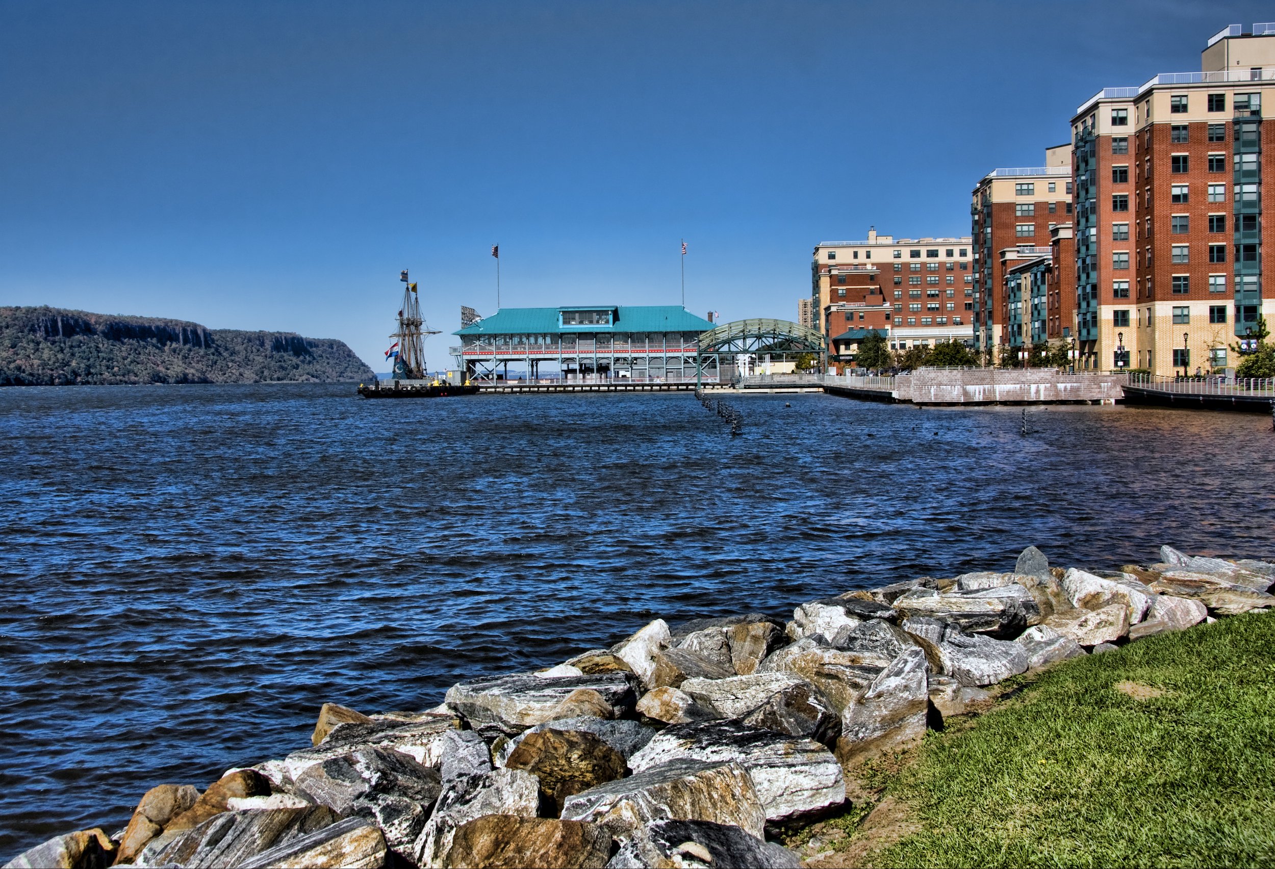 A waterfront view with a rocky shoreline in the foreground, a body of water, and a building pier with a blue roof and a ship replica, surrounded by residential buildings under a clear blue sky.