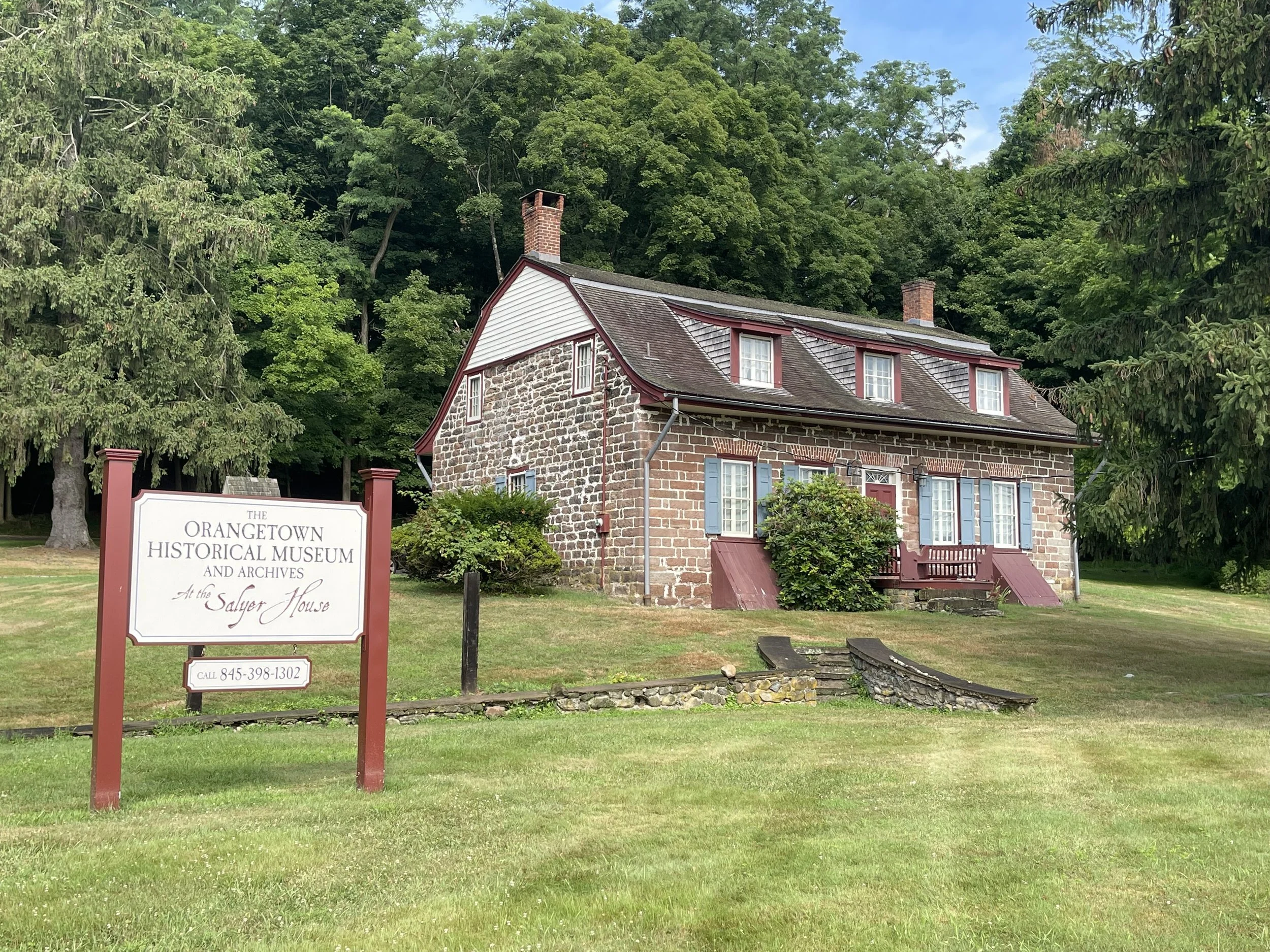 Photo of a historic stone house with a sign in front that reads 'The Orangetown Historical Museum and Archives at the Salyer House,' surrounded by trees and a well-kept lawn.