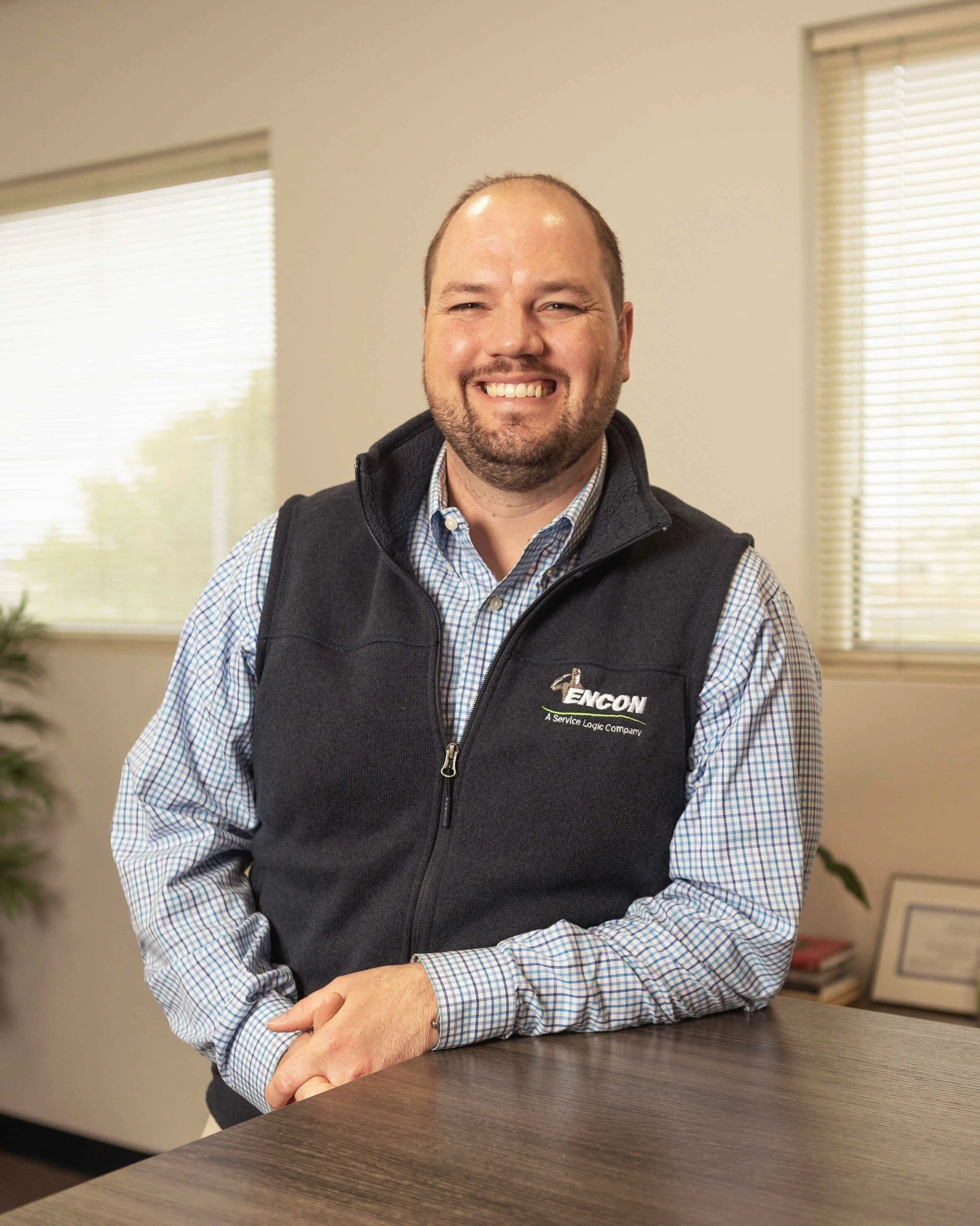 A smiling man with a beard and short hair standing indoors, wearing a checkered shirt and a black vest with the company logo 'Encon'.