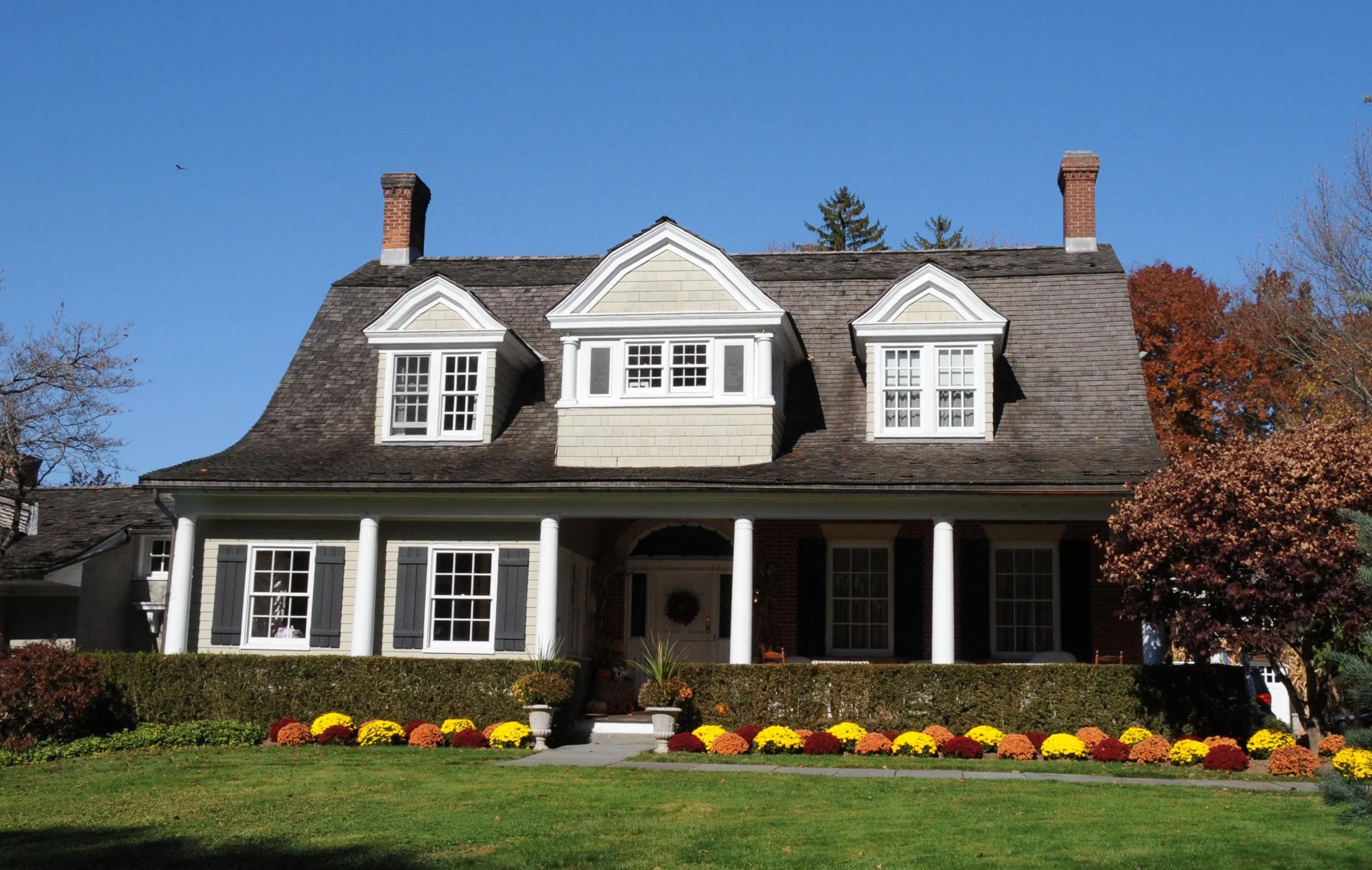 A two-story house with a large front porch, white columns, and a gabled roof. The house has multiple windows with white trim and black shutters. There are colorful flower beds with yellow, orange, and red flowers in front, and a well-manicured lawn. A red brick chimney is on each side of the roof, and the sky is clear and blue.