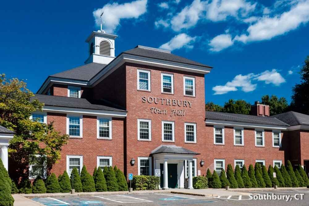 Southbury Town Hall building made of red brick with white-framed windows, a small central entrance with a porch, and a cupola on top, located in Southbury, Connecticut under a blue sky with clouds.
