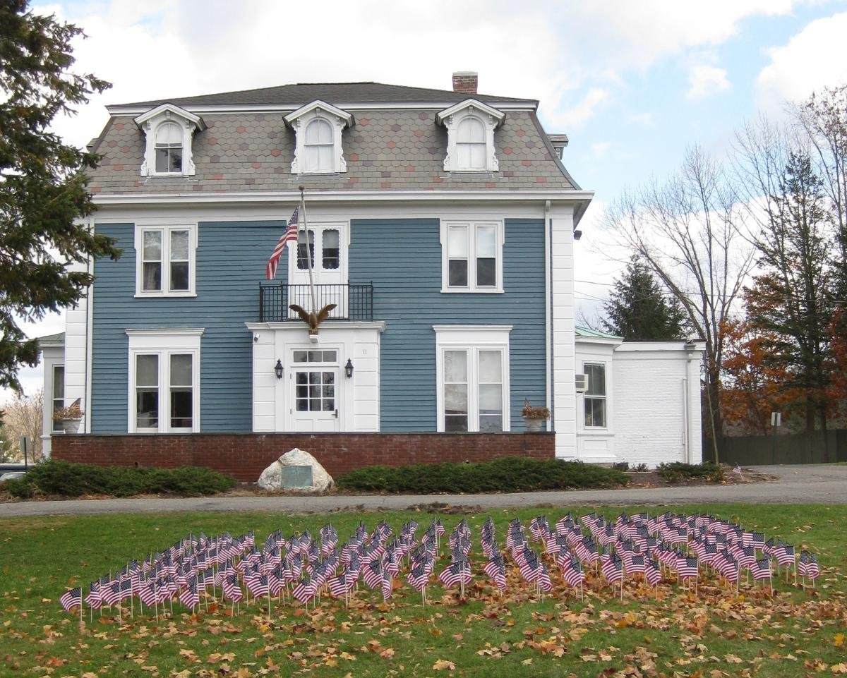 A large historical house with blue and white siding, multiple windows, small dormer windows on the roof, and a front porch. The yard displays numerous small American flags arranged in rows on the grass, with some fallen leaves, suggesting a patriotic display.