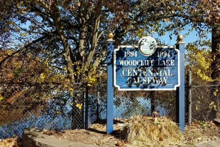 Blue sign commemorating the 100th anniversary of Woodcliff Lake Centennial Causeway, with a tree and a water background.