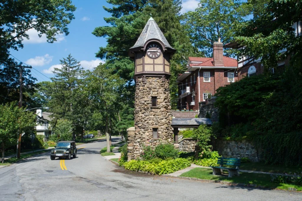 A stone clock tower on a residential street with houses and trees, blue sky with clouds.