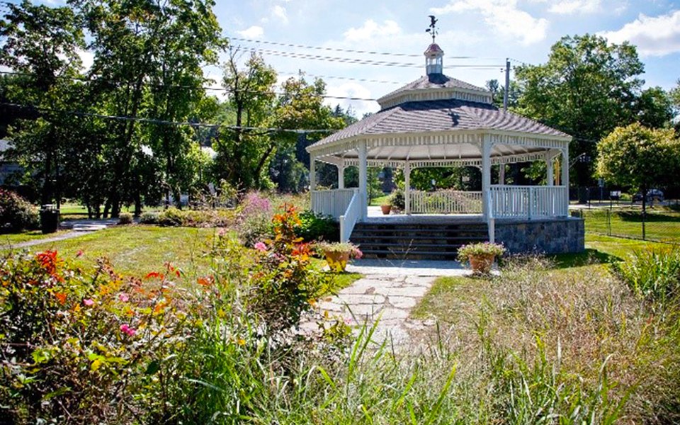 A white gazebo with stairs and a metal roof in a park, surrounded by colorful flowers and green trees, under a partly cloudy sky.