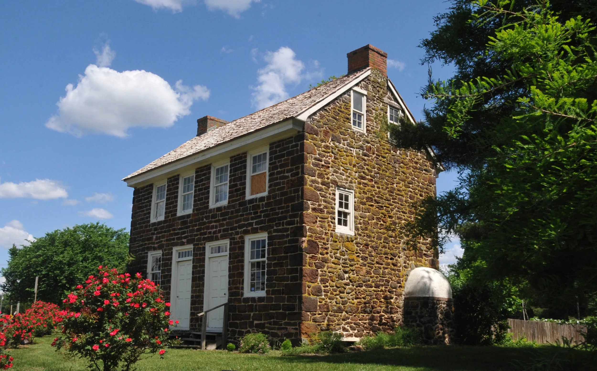 An old stone house with white trim, a steep shingled roof, and several windows. The house is surrounded by green trees and bushes, with pink flowers in the lawn. The sky is bright blue with a few white clouds.