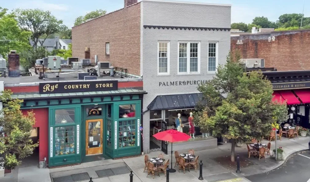Downtown street view featuring two storefronts, a small outdoor seating area with tables and red umbrellas, and a tree, with a brick building and other small structures in the background.