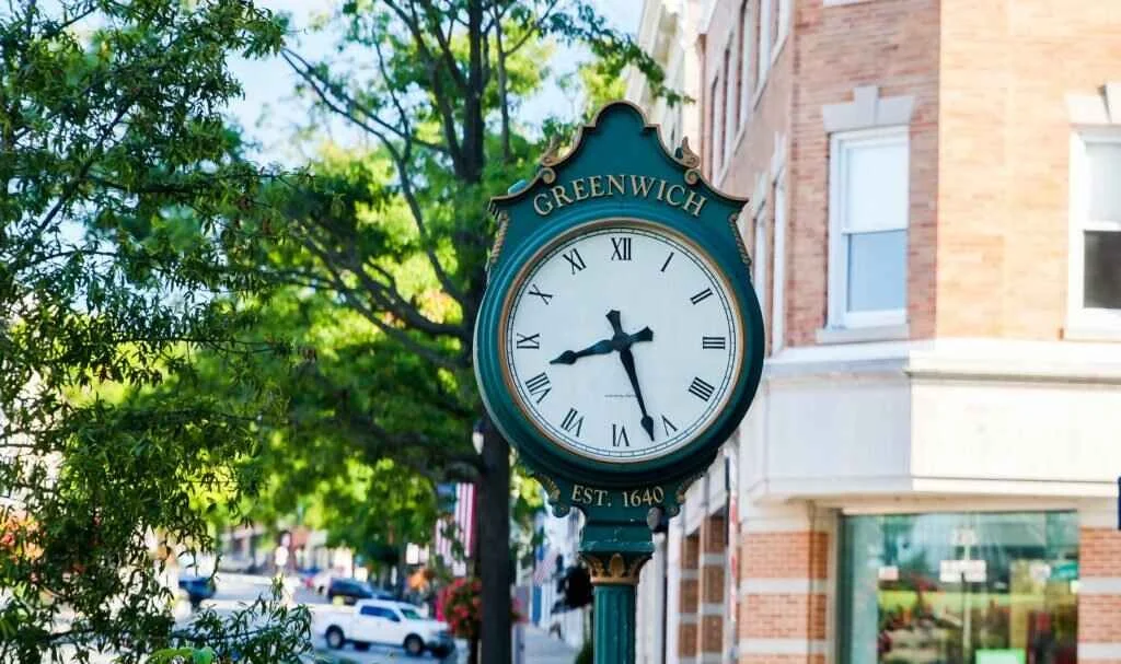 Street clock with green frame reading Greenwich, established 1640, in front of a brick building, with trees and parked cars in the background.