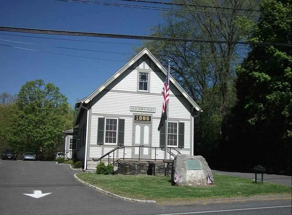 A small, white, wooden town hall building with a steep roof, located on a corner lot with a grassy area and a large stone monument in front. An American flag and a state flag are flying on a flagpole in front of the building. The sign above the door reads "Old Town Hall" and the year "1886" is displayed on the front.