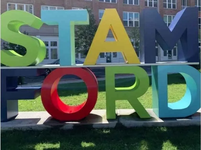 Colorful sculpture spelling out 'STAMFORD' with each letter in a different bright color, located outdoors on a grassy area with residential buildings in the background.